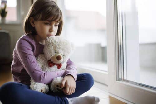 Sad lonely little girl hugging her teddy bear by the window